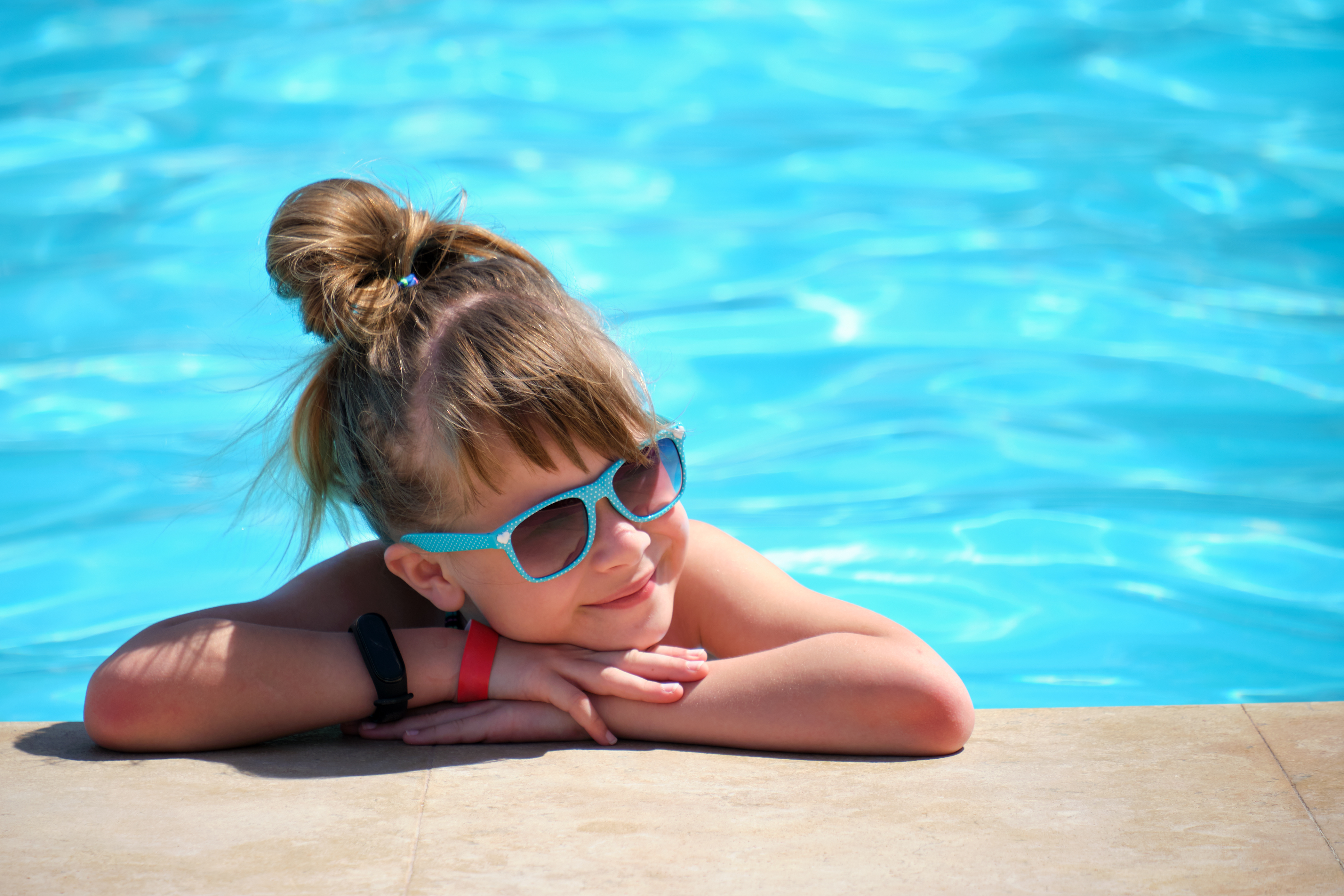 Young joyful child girl resting on swimming pool side with clear blue water on sunny summer day. Tropical vacations concept.